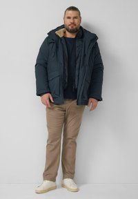 Steppjacke in Dunkelblau mit weichem beigem Futter, Reißverschluss und Druckknopfverschlüssen, mehrere Taschen. Beige Hose und weiße Turnschuhe runden das Outfit ab.