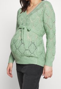 Pull de maternité vert tricoté avec col en V, motif en losange et ceinture à la taille. Il possède des manches longues et des poignets côtelés. Finition texturée.