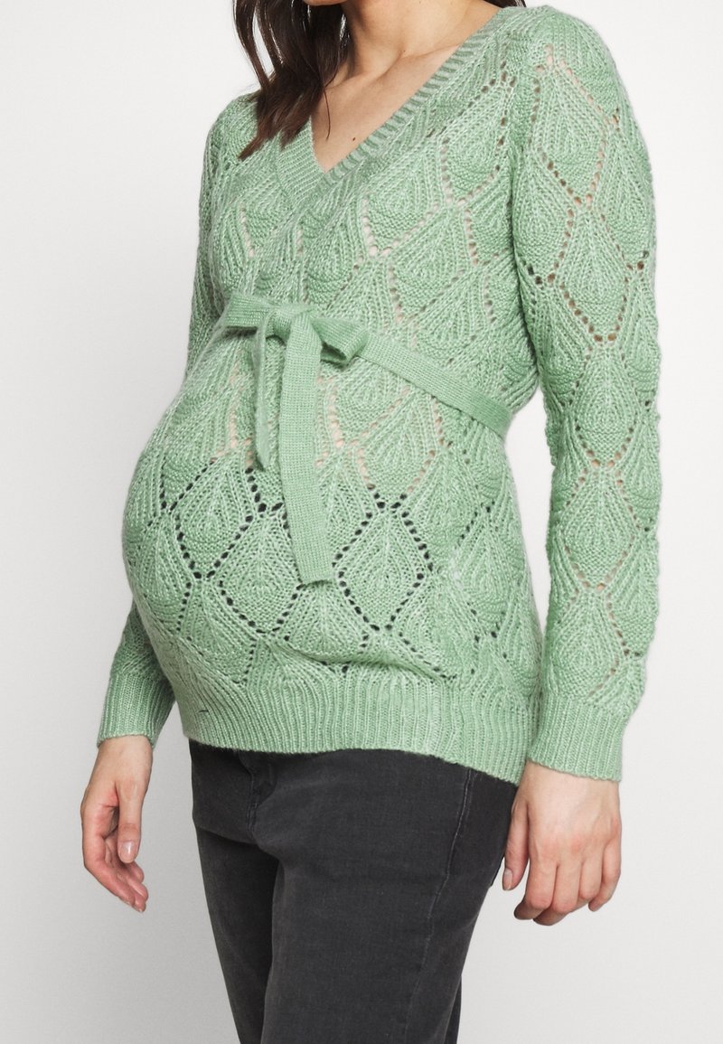 Pull de maternité vert tricoté avec col en V, motif en losange et ceinture à la taille. Il possède des manches longues et des poignets côtelés. Finition texturée.