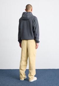 Grå fleecetröja med huva, kombinerad med gula vida byxor. Tyget verkar mjukt och outfiten har minimal detaljer.