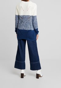 Stickad tröja med tre horisontella färgblock: krämfärgad, ljusblå och mörkblå. Löst snitt med öppet vävda mönster på baksidan. Matchas med mörka denim-byxor med vida ben.