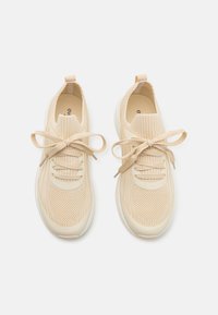 Beige strikkede joggesko med en teksturert mesh-overdel, polstret krage og matchende snøring. Hvit gummisåle og subtil logo på innersålen.