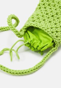 Borsa in macramè intrecciata verde chiaro con fodera in tessuto a coulisse e lungo manico intrecciato su sfondo bianco.