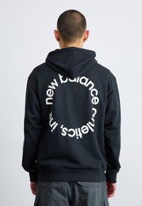 Haut à capuche noir avec un cordon de serrage et un motif circulaire blanc imprimé au dos, affichant le texte "new balance" dans une police moderne.