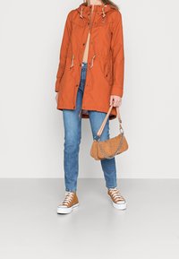 Pomarańczowa kurtka parka z kapturem i regulowanymi sznurkami, z przednimi kieszeniami i zatrzaskami. Połączona z niebieskimi dżinsami i brązowymi butami sportowymi, trzyma torbę w kolorze brązowym.
