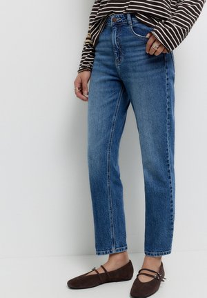 Kvinde iført blå jeans med høj talje, brune flade sko med dobbelt strop, og en langærmet brun- og hvidstribet skjorte med ringe på fingrene.