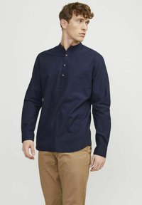 Camicia a maniche lunghe blu navy con colletto alla coreana, caratterizzata da una pattina con bottoni e una texture sottile. Abbinata a pantaloni beige.