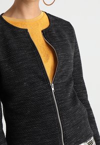 Giacca nera in tessuto strutturato con chiusura a zip e scollo rotondo. Indossata sopra un top giallo, con dettagli eleganti e hardware in metallo argentato.