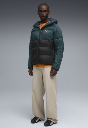 Geplüschte Jacke in Teal und Schwarz, mit einem Reißverschluss auf der Vorderseite, Kapuze und Seitentaschen. Kombiniert mit einem orangefarbenen Hemd und beigen Hosen.