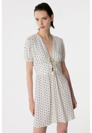 Giovane donna che indossa un vestito bianco a maniche corte con pois neri e dettagli con lacci sul davanti, in piedi davanti a uno sfondo semplice.