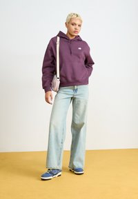 Lila puuvillane kapuutsiga särk koos ees oleva taskuga, koos helesiniste laia jala teksade ja siniste spordijalatsitega, seistes kollasel põrandal.