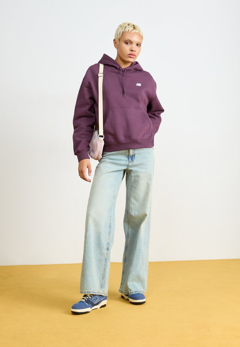 Haut à capuche en coton violet avec poche avant, associé à un jean large bleu clair et des baskets bleues, debout sur un sol jaune.