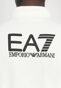 Bela polo majica s velikim črnimi vezenimi logotipom na hrbtu, z napisom "EA7" in "EMPORIO ARMANI" v kontrastnih stilih pisave.