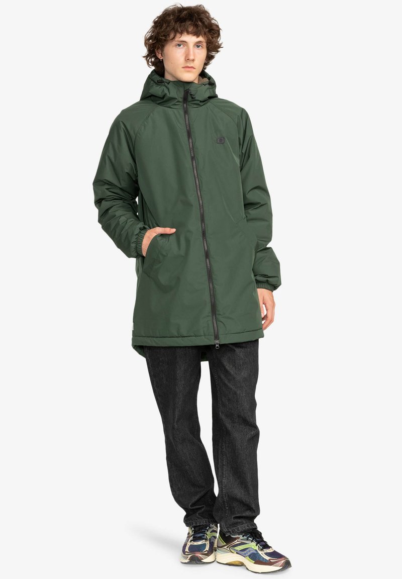 Giacca impermeabile verde con cappuccio, chiusura a zip, polsini elasticizzati e due tasche laterali, abbinata a jeans scuri e sneakers multicolori.