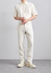 Kremowa koszula z krótkim rękawem o fakturowanej tkaninie, zestawiona z jasnymi lnianymi spodniami i beżowymi wsuwanymi butami z grubą podeszwą.