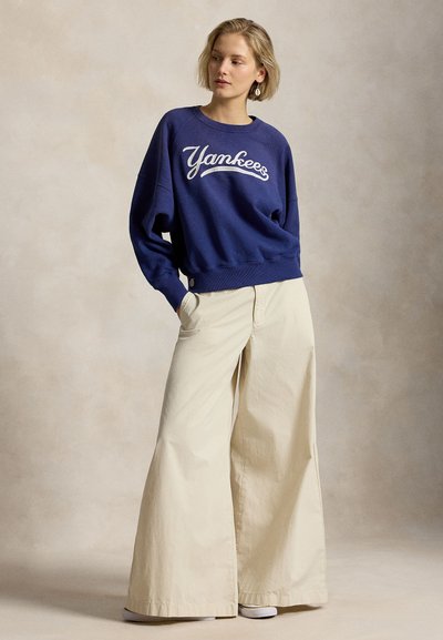 Νεαρή γυναίκα με κοντά ξανθά μαλλιά που φοράει σκούρο μπλε φούτερ "Yankees" και εκρού φαρδιά παντελόνια, στέκεται με τα χέρια στις τσέπες.