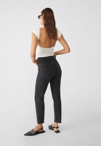 Mujer con cabello largo castaño, vistiendo una blusa blanca de espalda descubierta, jeans oscuros de tiro alto, zapatos planos negros de punta y gafas de sol, de pie frente a un fondo blanco.