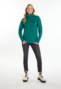 Zöldeskék kabelmintás garbó pulóver hosszú ujjal, fekete slim-fit farmernadrággal és krémszínű harci csizmával, fekete talppal párosítva.