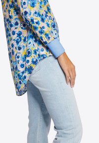 Bloemige blouse met blauwe, gele en roze bloemen, voorzien van lange mouwen en een lichtblauwe manchette. Gecombineerd met lichtblauwe spijkerbroek.