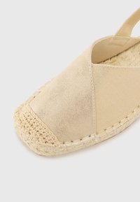 Bež espadrila s teksturiranim platnenim telom. Ima okrugli vrh, tkanu jutu kao đon i elastične trake za sigurno prianjanje.