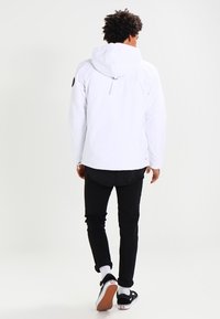 Chaqueta blanca con capucha de corte holgado, llevada sobre pantalones negros y zapatillas. Cuenta con un cordón ajustable en la espalda y un logo en la manga.