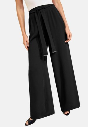 Vrouw die zwarte wijde broek met hoge taille draagt met een bijpassende stoffen riem, gecombineerd met zwarte open-toe hakken en een witte mouwloze top.