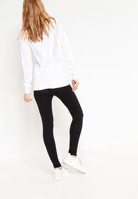 Weißes Langarmoberteil mit durchbrochenem Rückendesign, kombiniert mit schwarzen Leggings und weißen Sneakers, die kontrastierende Farben und Materialien hervorheben.