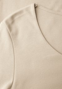 Beżowy materiał o gładkiej fakturze, z okrągłym dekoltem i krótkimi rękawami, z delikatnym streczem. Brak widocznych wzorów lub elementów metalowych.