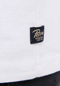 Bílá bavlněná tričko s našitým lemem a námořnickou modrou etiketou s nápisem "Petrol Industries Since 1983" ve zlatém písmu.