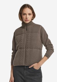 Brauner Zip-Hoodie mit hohem Kragen, versehen mit Kabelstrickmustern und langen Ärmeln. Weiche Textur und figurbetontes Design.