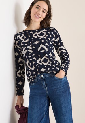 Donna appoggiata a un muro, che indossa un top lungo a maniche con un motivo geometrico blu navy e beige e jeans blu, mentre tiene un indumento in bordeaux piegato.