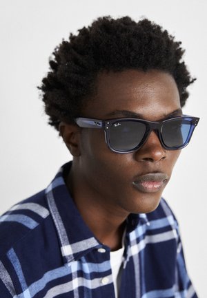 Blaue Ray-Ban Sonnenbrillen mit glänzender Oberfläche, quadratischer Form und dunklen Gläsern; getragen mit einem karierte Hemd in Marineblau und Hellblau.