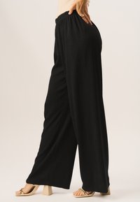 Black wide-leg pants with textured fabric, featuring an elastic waistband. The model wears gold strappy heels.