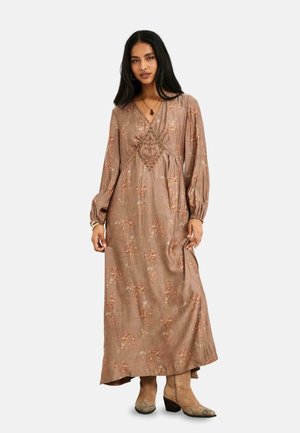 Mujer con cabello largo y oscuro que lleva un vestido largo marrón con flores y mangas largas, y botas cortas color cuero claro, de pie frente a un fondo liso.