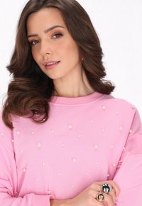 Felpa rosa con decorazioni di perle sparse, scollatura rotonda e maniche lunghe. Il modello mostra un primo piano del top e degli accessori.