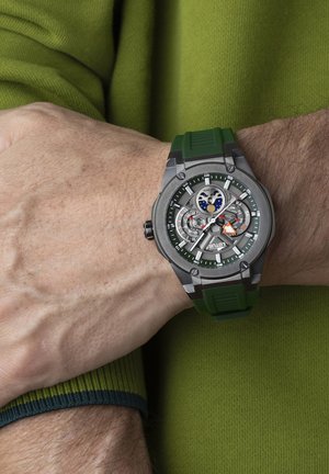 Montre avec un bracelet en silicone vert et un boîtier en métal gris, cadran complexe avec trois sous-cadrans et accents colorés, affichée sur un poignet.