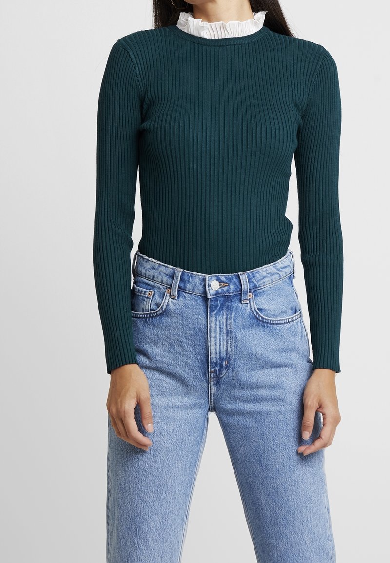 Femme portant un haut côtelé vert foncé à manches longues avec un col blanc à volants, associé à un jean bleu clair taille haute.