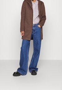 Cappotto marrone con cappuccio sopra una maglietta bianca e viola a righe, abbinata a jeans larghi blu e scarpe chunky nere.