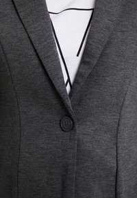 Nærbillede af en mørkegrå blazer med en enkelt sort knap, båret over en hvid skjorte med et sort geometrisk design.