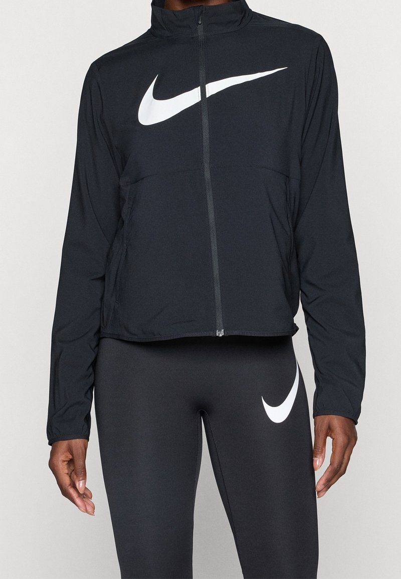 Personne portant une veste zippée Nike noire et des leggings noirs avec des logos Nike Swoosh blancs sur la poitrine et la cuisse.