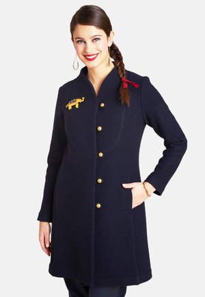 Marineblauer Wollmantel mit goldenen Knöpfen und einem dekorativen Elefantenaufnäher. Gerader Schnitt, zwei Seitentaschen und hoher Kragen.