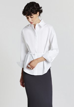 Donna con capelli corti e ricci, che indossa una camicia bianca con cintura e una gonna textured scura, guarda in basso con la mano appoggiata sui fianchi.