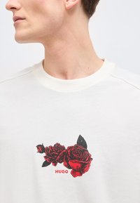 Άνδρας που φοράει λευκό μπλουζάκι με στρογγυλή λαιμόκοψη, με γραφικό κόκκινο και μαύρο τριαντάφυλλο και το κείμενο "HUGO" στο στήθος, κοιτάζει αριστερά με ορατό λαιμό και σαγόνι.