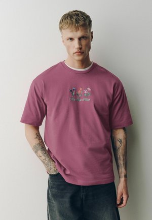 Junger Mann mit kurzen blonden Haaren, der ein lila T-Shirt mit Blumenstickerei und schwarze Jeans trägt, steht vor einer schlichten weißen Wand.