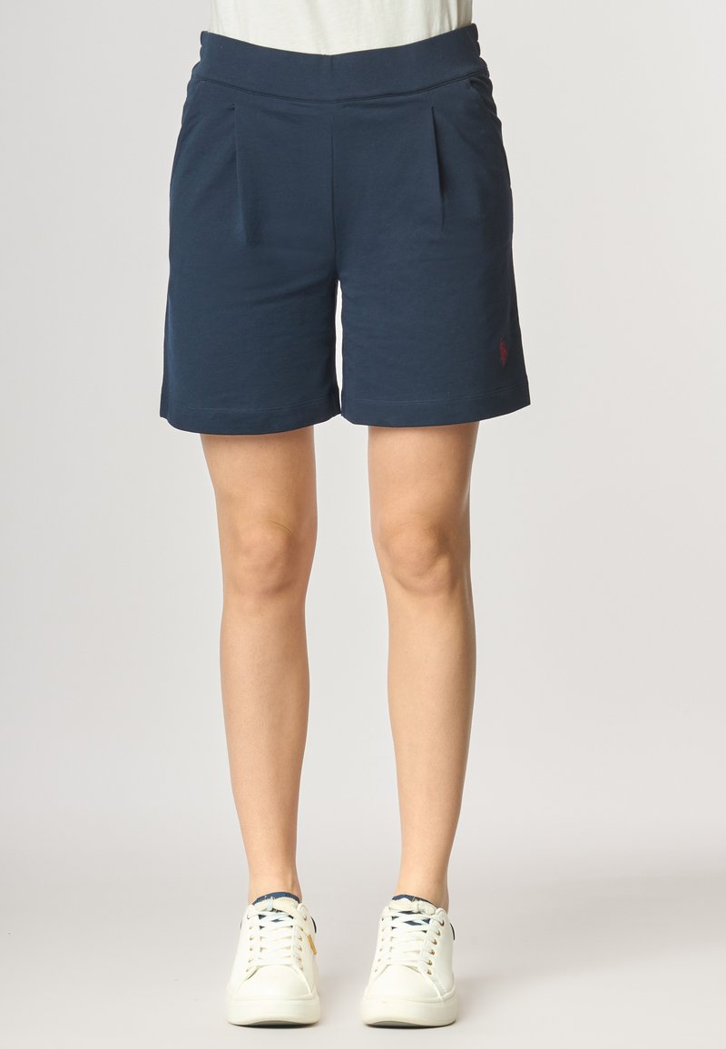Pantaloni corti blu navy con una texture liscia, presentano una cintura ampia, due pieghe frontali e un piccolo dettaglio con logo rosso sul lato inferiore destro.
