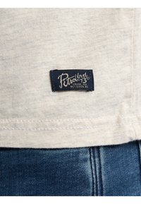 Camiseta de algodón gris claro con etiqueta azul marino con costuras blancas; la etiqueta dice "Petrol 73 Crude Oil" con textura de tela visible.