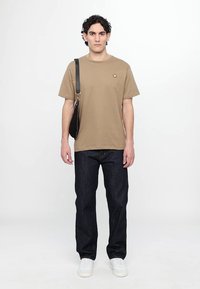 Beige Baumwoll-T-Shirt mit Rundhalsausschnitt; blaue Jeans; weiße Sneakers; schwarze Umhängetasche mit Kettenriemen.
