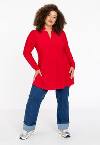 Rode tuniek met lange mouwen en een V-hals, licht uitlopende snit, gecombineerd met omgeslagen blauwe denim jeans en zilveren sneakers.