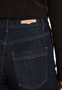 Dark wash denim jeans with a small leather tag at the waistband. Features orange stitching and two back pockets.
