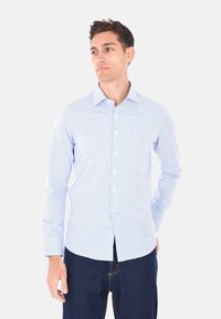 Camicia azzurra chiara con strisce verticali bianche, maniche lunghe, colletto classico e orlo dritto. Abbinata a jeans di denim scuro.